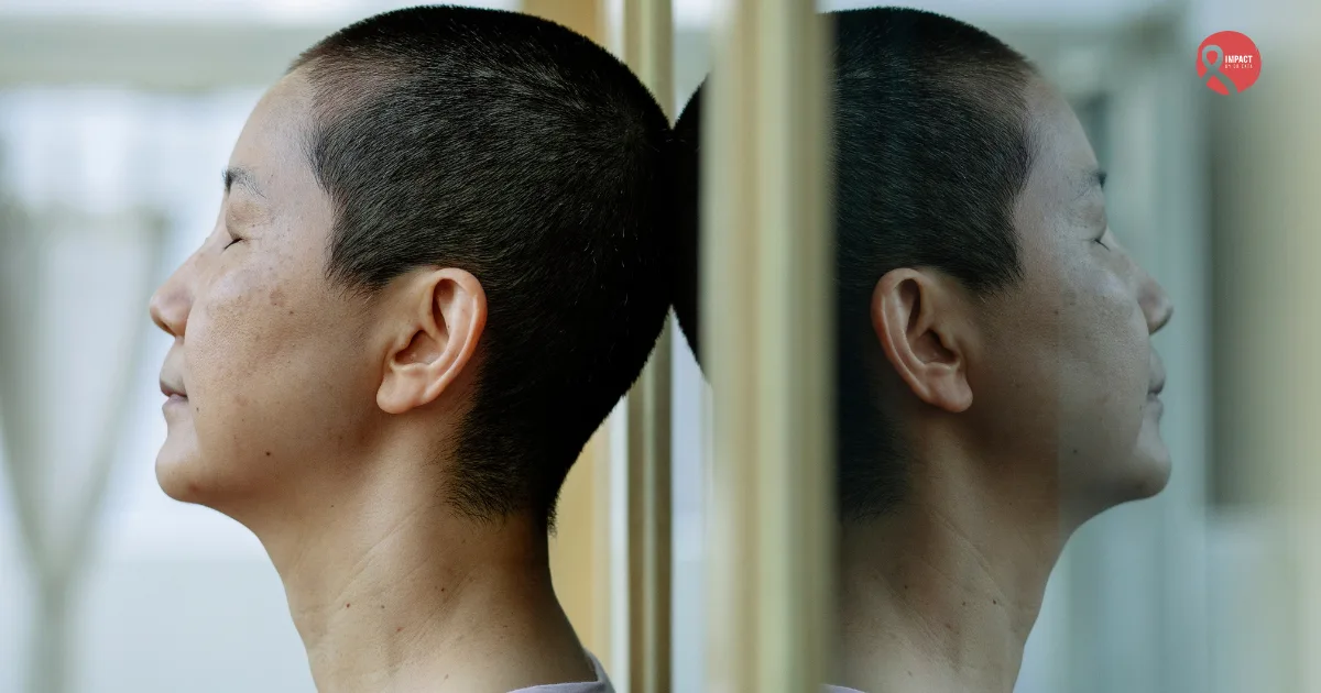 A cancer patient with a shaved head rests against a glass wall, reflecting on work-life balance during treatment.